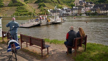 Radfahrer überqueren bei Beilstein die Mosel mit einer Fähre, während Menschen am Ufer dabei auf einer Bank sitzen.  | Bild: dpa-Bildfunk/Thomas Frey