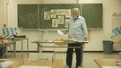 Älterer Lehrer, welcher vor seiner Tafel im Klassenzimmer steht. Auf der Tafel sind mehrere Objekte mit der Bairischen Bezeichnung zu sehen. | Bild: BR