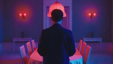 Ein Mann im Anzug, von hinten gefilmt, welcher vor einem Neon belichteten Blau-Pinken Zimmer steht. Vor ihm steht ein Esstisch mit Stühlen. | Bild: BR