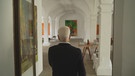 Ein äterer Mann, welcher am Eingang von einem großen, komplett in weiß gehaltenen Atelier steht. Im Hintergrund hängen mehrere Gemälde. | Bild: BR