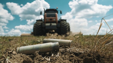"DokThema" geht der Frage nach, wie es so weit kommen konnte, dass Weizen als politisches Druckmittel eingesetzt werden kann. Granathülsen, die auf einem Acker in der Nähe von Mykolajiw liegen. Im Hintergrund ein Traktor. | Bild: BR/WDR