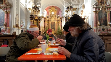 Menschen sitzen in einer Kirche am Tisch und essen Suppe | Bild: BR