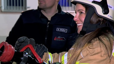 Reporterin in Feuerwehr-Montur mit Wasserschlauch in der Hand. | Bild: BR