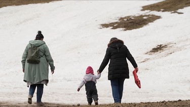 Zwei Frauen und ein Kind laufen auf einer Wiese mit Schnee-Resten. | Bild: BR