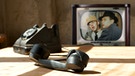 Antikes Telefon auf einem Tisch, im Hintergrund ein Fernseher auf dem ein Szenenbild aus der "Löwengrube" zu sehen ist | Bild: colourbox.com, BR/Tellux-Film GmbH; Montage: BR
