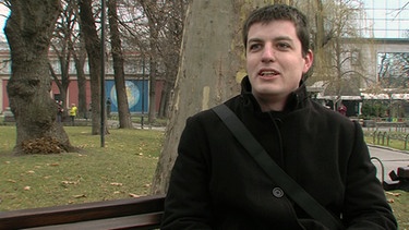 Ein junger Mann sitzt auf einer Parkbank und spricht in die Kamera | Bild: BR