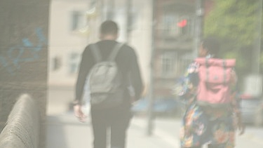 In einer milchig eingetrübten Szene sieht man zwei Menschen mit Migrationshintergrund, die auf einem Bürgersteig nebeneinander gehen. | Bild: BR