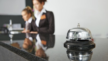 Eine Rezeptionsglocke in einem Hotel. Im Hintergrund zwei Mitarbeiterinnen des Hotels. | Bild: stock.adobe.com/Kadmy