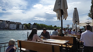 Blick auf ein Café mit Menschen, die auf Bänken sitzen. Das Café liegt direkt am Bodensee. | Bild: Wir in Bayern