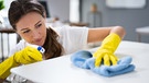 Eine Frau trägt gelbe Handschuhen und reinigt mit einem Tuch eine Tischfläche. | Bild: stock.adobe.com/Andrey Popov