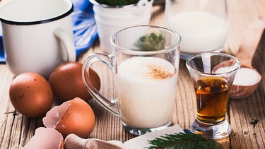 Eine Glastasse gefüllt mit einer Mischung aus Milch und Sahne, links davon liegen zwei ganze Eier und eine aufgeschlagene Eierschale, rechts davon ein Schnapsglas mit Doppelkorn. Dahinter ist eine Glaskaraffe mit Milch und einem Milchtopf zu sehen. Alles steht auf einem braunen Holztisch. | Bild: mauritius images / Istetiana / Alamy