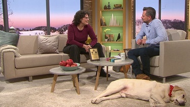 Moderator Michael Sporer spricht mit Gast Christine Thürmer, der "Langstreckenwanderin", Hund Henry liegt am Boden | Bild: BR / "Wir in Bayern"