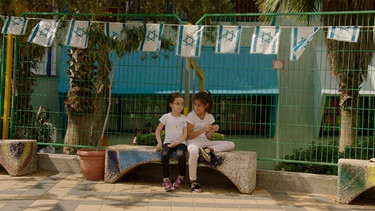 Zwei Mädchen auf einer Steinbank vor einem Zaun an einer Grundschule in Petach Tikva. | Bild: ZDF/WDR/Andreas Köhler