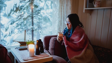 Eine  Frau hüllt sich in eine wärmende Decke, draußen schneit es. | Bild: picture alliance / Shotshop / Addictive Stock