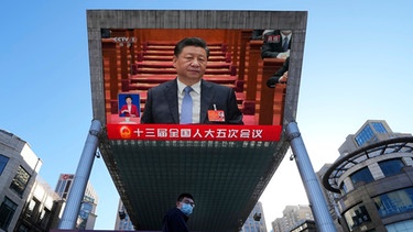 05.03.2022, Peking: Eine Person geht in einem Einkaufszentrum an einem großen Videobildschirm vorbei, auf dem die Rede des chinesischen Präsidenten Xi Jinping während der Eröffnungssitzung der Jahrestagung des Nationalen Volkskongresses (NVK) live übertragen wird. | Bild: dpa-Bildfunk/Ng Han Guan
