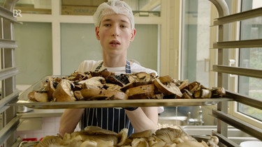 Simon macht eine Ausbildung und schiebt ein Backblech mit Pilzen in einen Ofen. | Bild: Bayerischer Rundfunk 2024
