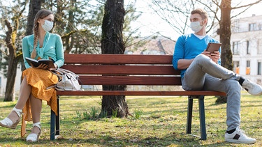Eine Frau und ein Mann sitzen mit großem Abstand und Masken auf einer Parkbank. | Bild: colourbox.com