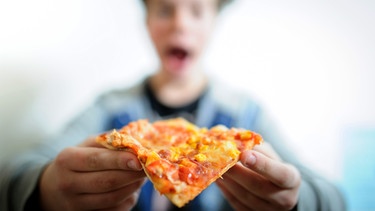 Ein Junge (verschwommen) isst ein Stück Tiefkühlpizza. | Bild: picture-alliance/dpa