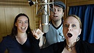 Zwei Frauen und ein Mann stehen im Studio vor einem Mikro mit offenem Mund. | Bild: BR