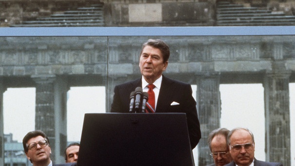 [12.06.1987] Der damalige US-Präsident Ronald Reagan redet vor der Berliner Mauer am Brandenburger Tor (Archivfoto vom 12.06.1987). Rechts sitzt der damalige Bundeskanzler Helmut Kohl (CDU), links der damalige Bundestagspräsident Philipp Jenninger (CDU). | Bild: picture-alliance/dpa