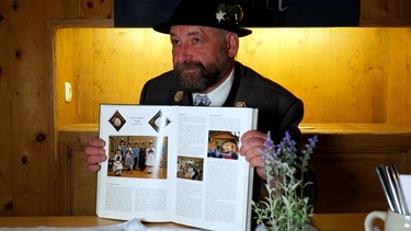 Der zuständige Gauvorstand, Michael Hauser.  Er trägt Trachtenhut, sitzt an einem Tisch und hält ein aufgeklapptes Buch in den Händen. | Bild: BR 