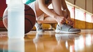 Sporthallenboden mit einem Ball und einem Ausschnitt wie sich jemand einen Turnschuh zuschnürt | Bild: picture alliance / Zoonar | Channel Partners