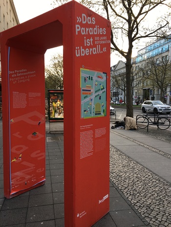 Ein Tor mit der Aufschrift "'Das Paradies ist überall.' 500 Jahre Reformation" steht auf dem Kurfürstendamm in Berlin am 22.04.2017 und weist auf den kommenden Kirchentag hin. Dahinter sitzt ein Bettler am Straßenrand. | Bild: Gerhard Brack
