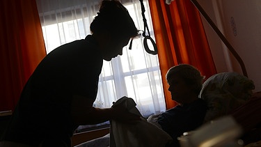 Eine Frau wird in ihrem Bett in ihrem Zimmer von einer Pflegerin betreut. | Bild: picture-alliance/dpa/Jens Kalaene