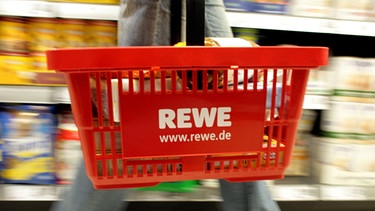 Eine Kundin mit einem tragbaren Plastikkorb mit REWE-Logo am Arm kauft ihre Lebensmittel in einem REWE-Supermarkt in Köln ein | Bild: picture-alliance/dpa