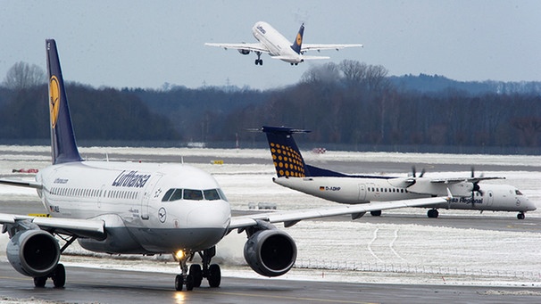 Zwei Lufthansa-Flugzeuge warten am auf dem Rollfeld des Flughafen München auf ihre Startfreigabe, während im Hintergrund eine Maschine abhebt | Bild: picture-alliance/dpa