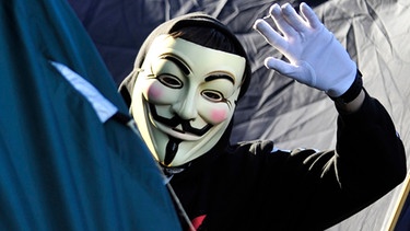 Ein Aktivist mit einer Guy-Fawkes-Maske, dem Erkennungszeichen der Hackergruppe Anonymous, winkt. | Bild: dpa/Marc Tirl