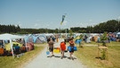 Drei Festivalgäste laufen einen Weg auf dem Campinggelände entlang. Sie tragen ihr Gepäck und eine Kühlbox. Neben ihnen sitzt bereits eine Gruppe unter einem Pavillon. Zahlreiche Zelte sind bereits aufgebaut.  | Bild: BR/Kilian Seiler