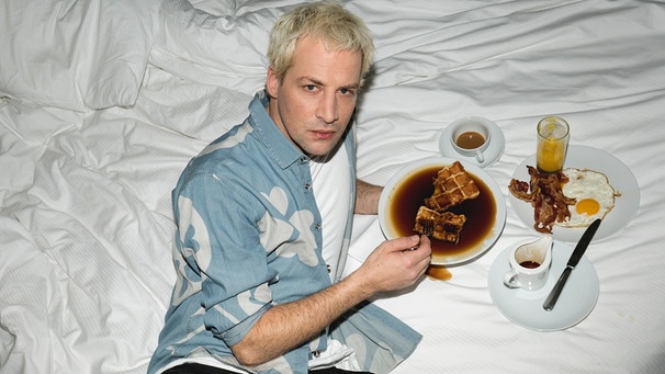 Rapper Dendemann liegt auf dem Bett und isst eine Waffel mit viel zu viel Sirup | Bild: Nils Müller