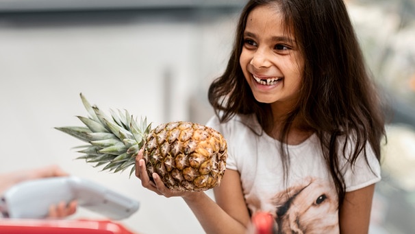 Ein kleines Mädchen freut sich beim Einkaufen über die Ananas in ihrer Hand | Bild: mauritius-images