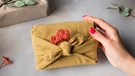 Eine weibliche Hand mit rot lackierten Fingernägeln hält ein Geschenk, dass in einem Tuch verpackt ist | Bild: mauritius-images