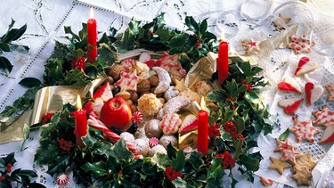 Ein Adventskranz aus Stechpalmenzweigen. In der Mitte liegen Plätzchen, Zuckerstangen und ein Apfel. Es brennen alle vier rote Kerzen. | Bild: mauritius images / ROSENFELD / Festlichkeiten / Celebrations