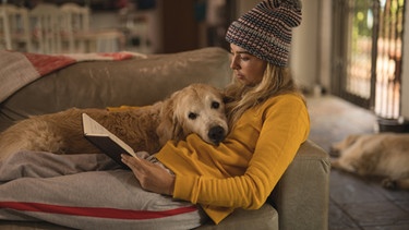 Frau liegt mit Hund und Buch auf der Couch | Bild: mauritius images / Wavebreakmedia