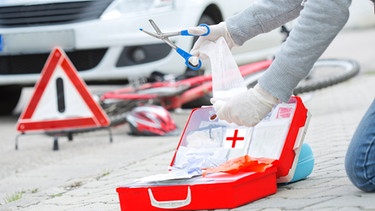 Jemand nimmt Dinge aus einem Erste Hilfe Kasten, dahinter Unfall | Bild: mauritius-images