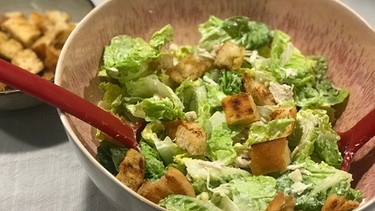 Eine Keramikschüssel mit Caesars Salad, garniert mit angeröstetem Weißbrot, steht auf einem Tisch | Bild: BR
