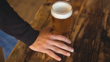 Ein Glas alkoholfreies Weißbier steht auf einem Tresen neben der Hand eines Mannes | Bild: mauritius images / Wavebreakmedia