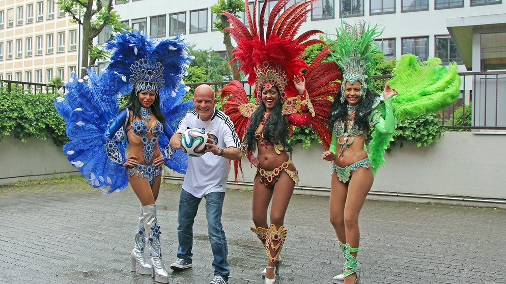 WM-Song: Bayern 1-Moderator Stephan Lehmann hält einen Fußball und wird von drei bunt geschmückten brasilianischen Tänzerinnen umringt | Bild: BR/Veronika Holzer