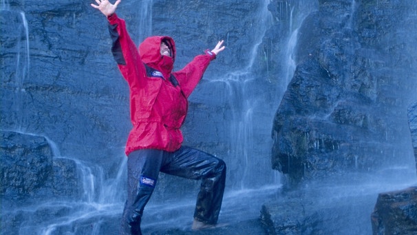 Ein Mann mit einer roten wasserabweisenden Jacke steht vor einem Wasserfall | Bild: mauritius-images