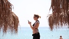 Eine Frau geht mit dem Handy in der Hand am Strand entlang. | Bild: picture-alliance/dpa
