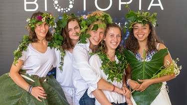 Fünf junge Frauen sind mit Grünpflanzen geschmückt und stehen vor dem Schriftzug "Biotopia". | Bild: Biotopia / Andreas Heddergott