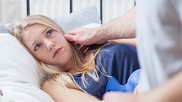 Frau im Bett mit kränklichem Blick, lässt sich von ihrem Partner trösten. | Bild: picture-alliance/dpa