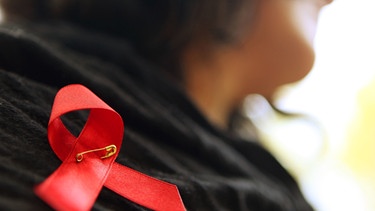 Eine Frau hat sich eine rote Schleife an den Kragen geheftet, Symbol der Solidarität mit an AIDS erkrankten oder mit HIV infizierten Menschen. | Bild: picture-alliance/dpa