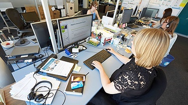Frau im Büro, zusammen mit anderen. Vor sich: Bildschirm, Kopfhörer, Smartphone, Büroutensilien | Bild: picture-alliance/dpa