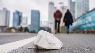 Eine weggeworfene FFP2-Maske liegt auf der Straße, im Hintergrund sieht man unscharf eine Stadt | Bild: dpa-Bildfunk/Frank Rumpenhorst