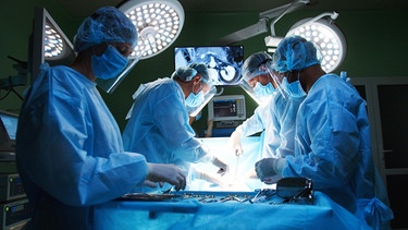 Ein Team aus Ärzten mit sterilen Kitteln, Handschuhen und OP-Hauben führen eine Operation in einem Operationssaal durch. | Bild: stock.adobe.com/ihorvsn