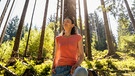 Eine Frau lehnt mit entspanntem Gesicht an einem Baum im Wald bei sonnigem Wetter. | Bild: picture alliance / Westend61 | Daniel Ingold